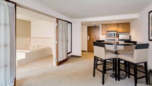 A modern hotel room with a bathroom, a dining area with a round table and chairs, and a kitchenette with a microwave and refrigerator.