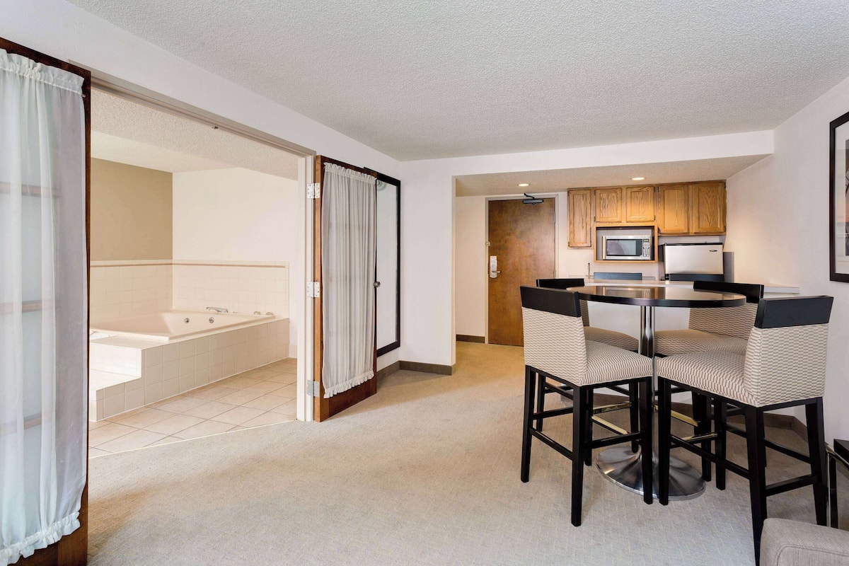 A modern hotel room with a bathroom, a dining area with a round table and chairs, and a kitchenette with a microwave and refrigerator.