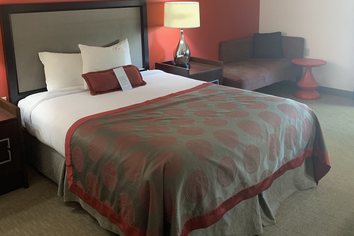 A hotel room with a bed, a nightstand, a lamp, a chair, and a small table.