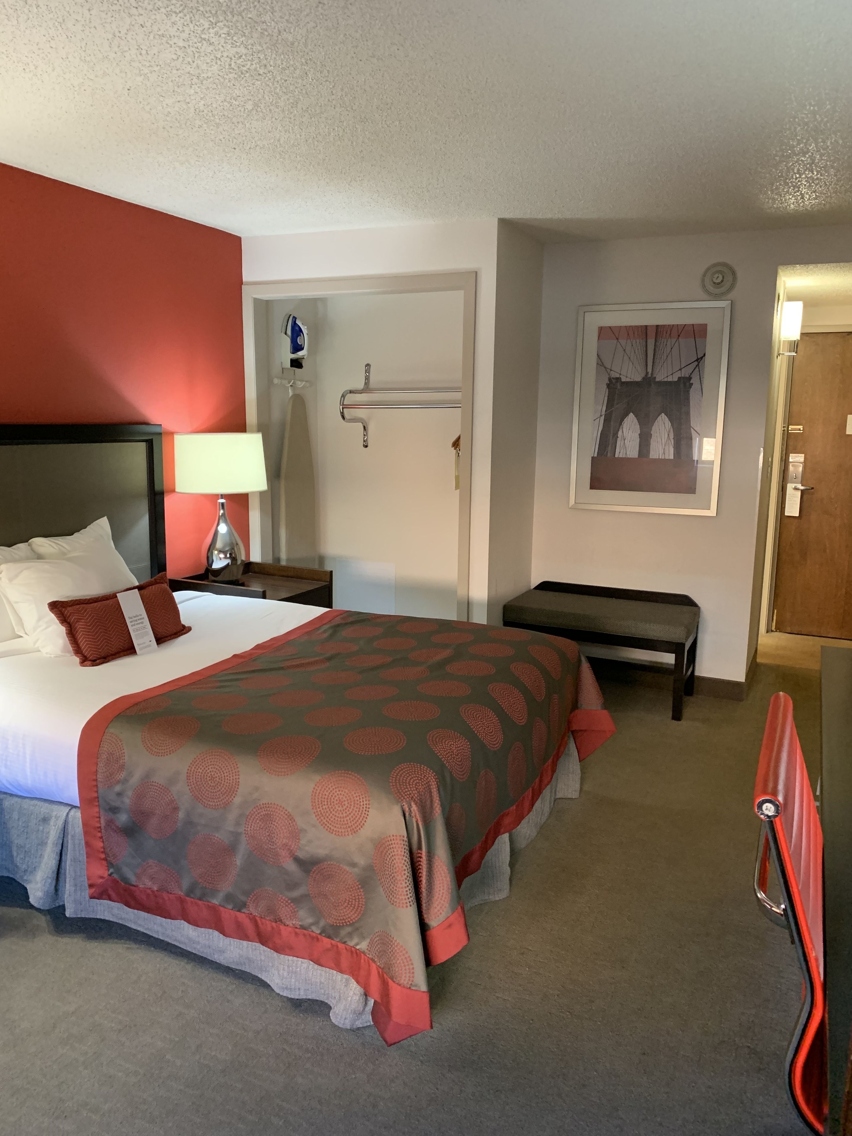 Quarto de hotel com cama, mesa de cabeceira, luminária, banco e uma porta.