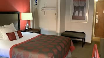 Een hotelkamer met een bed, nachtkastje, lamp, bankje en een deur.