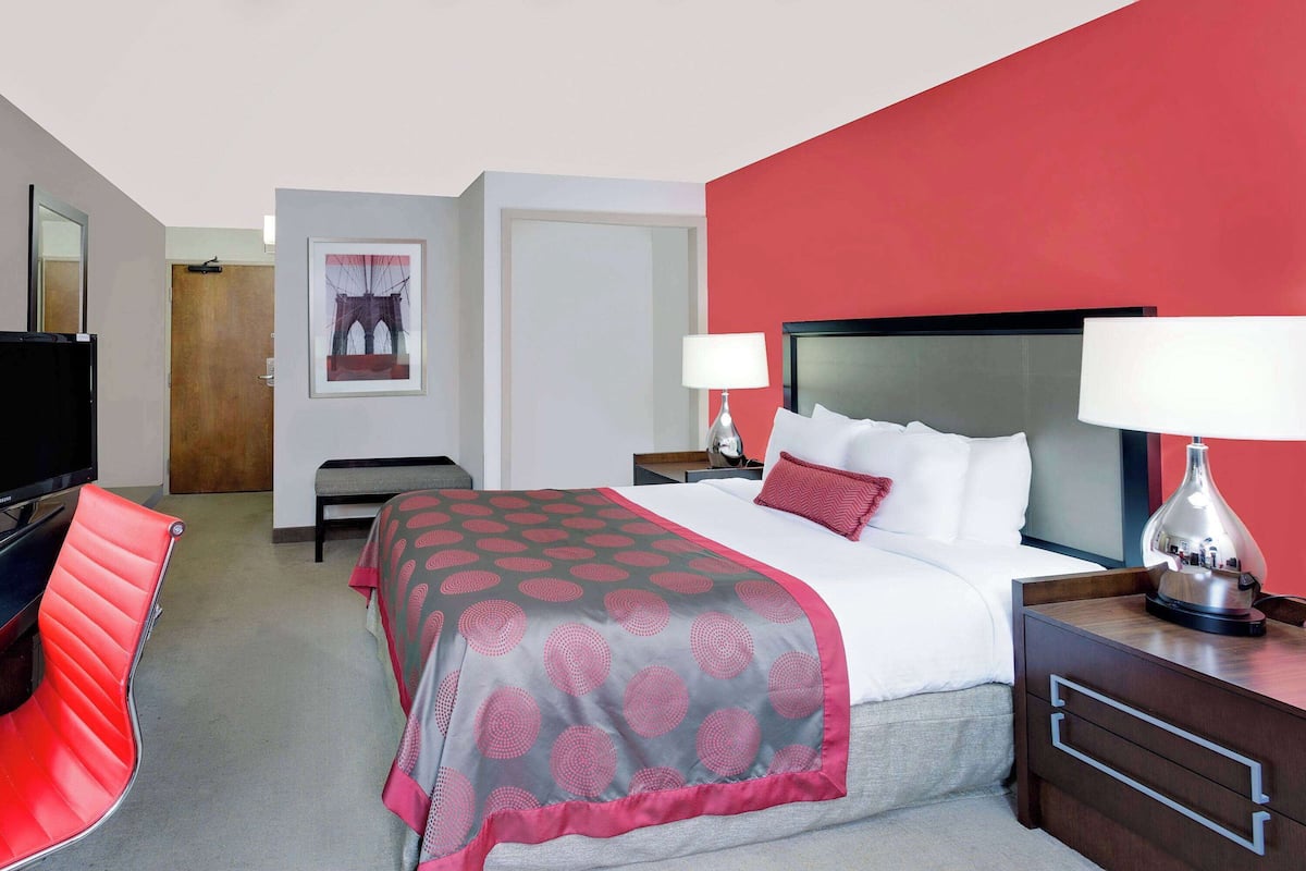 A hotel room with a bed, bedside tables, a TV, a red chair, and a painting on the wall.