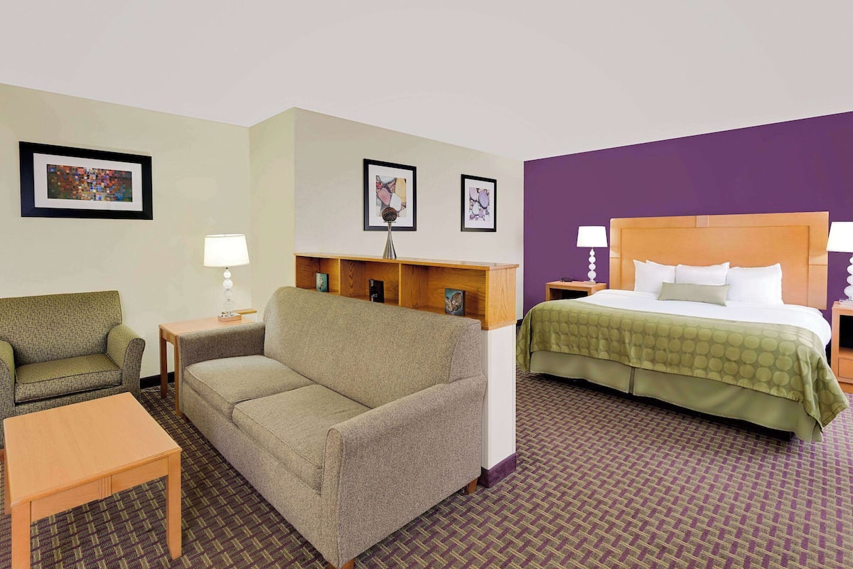 A hotel room with a sofa, a small table, a bed with a green bedspread, and two framed pictures on the wall.
