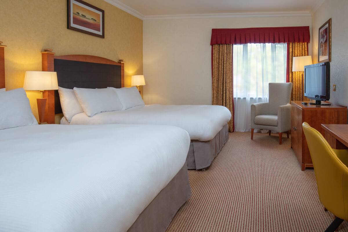 A hotel room with two beds, a desk with a television, a chair, and a lamp.