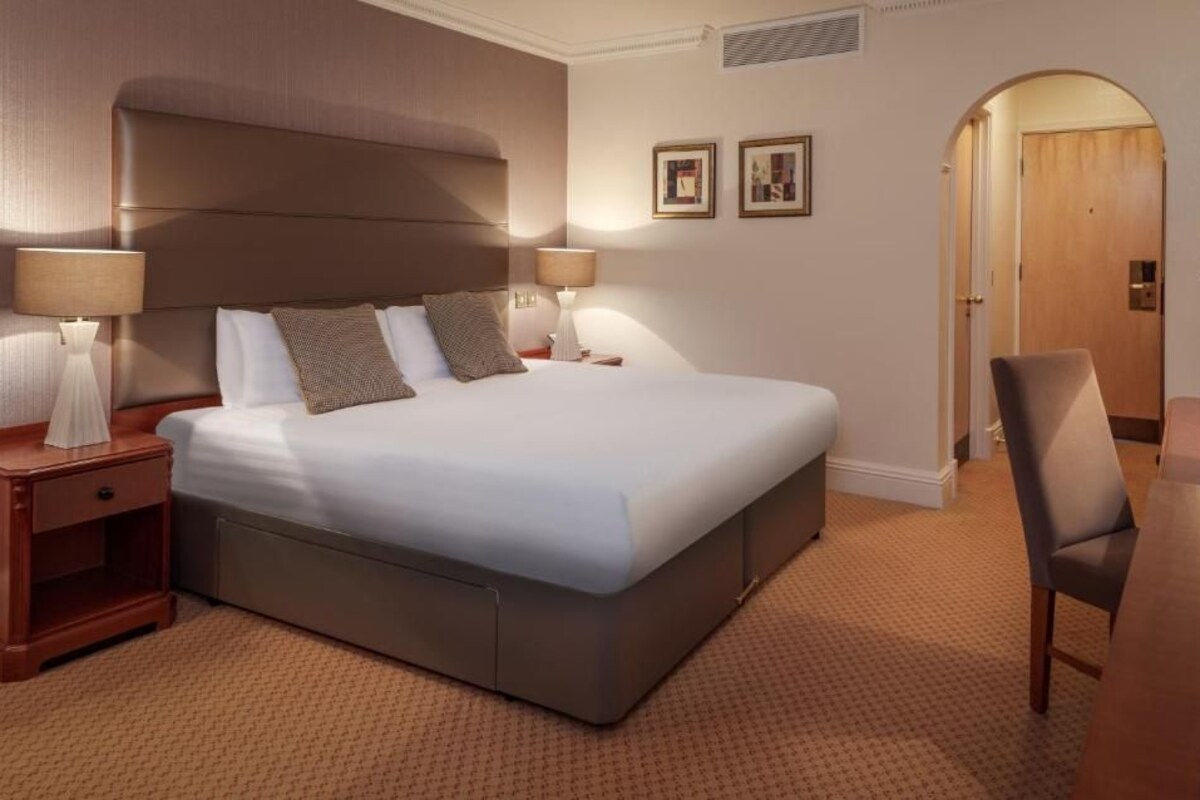 Una habitación de hotel con una cama grande, dos mesitas de noche con lámparas, una silla y una puerta que da a otra habitación.
