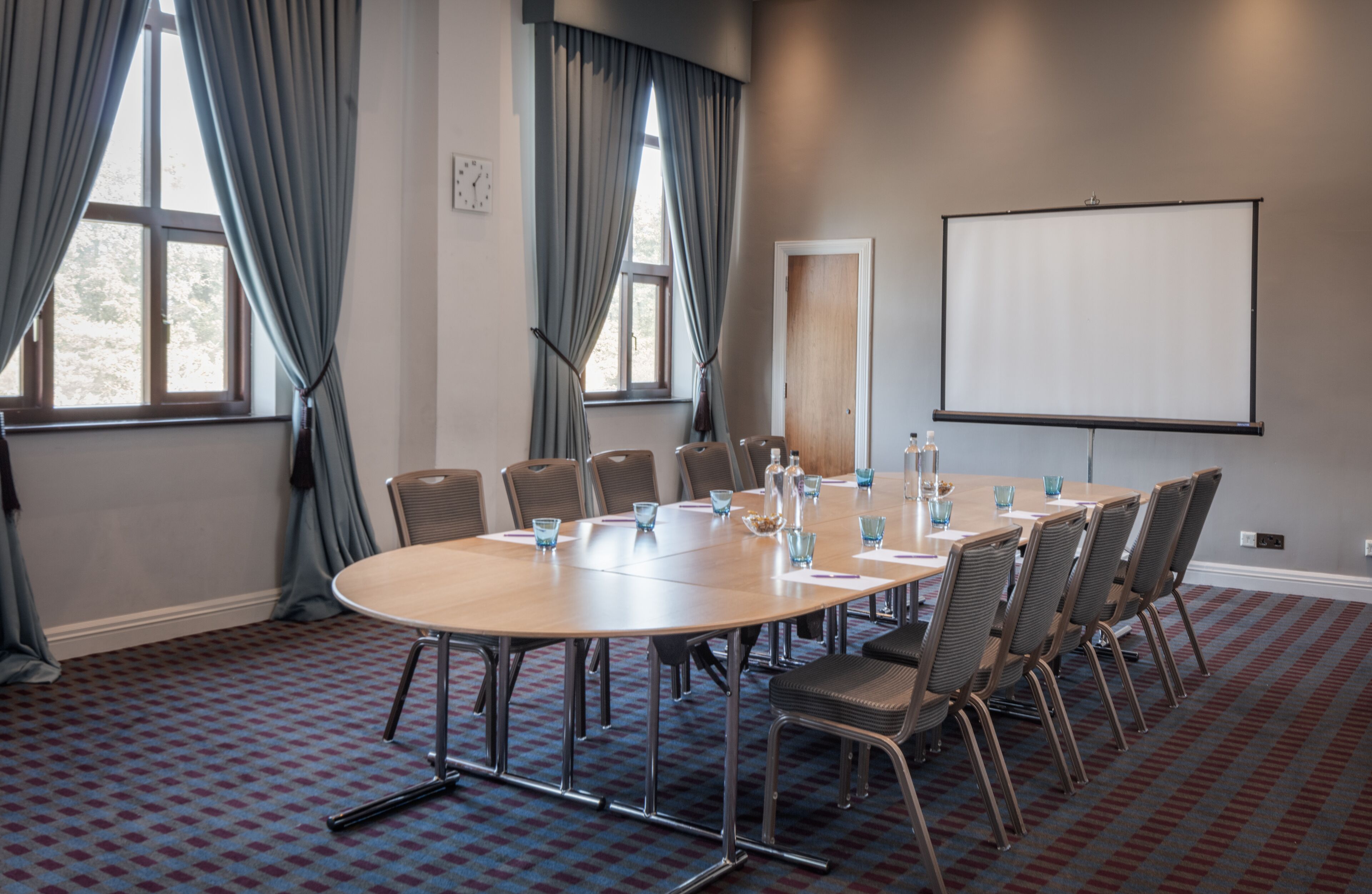 A conference room with a large oval table, chairs, a projector screen, and a clock on the wall.
