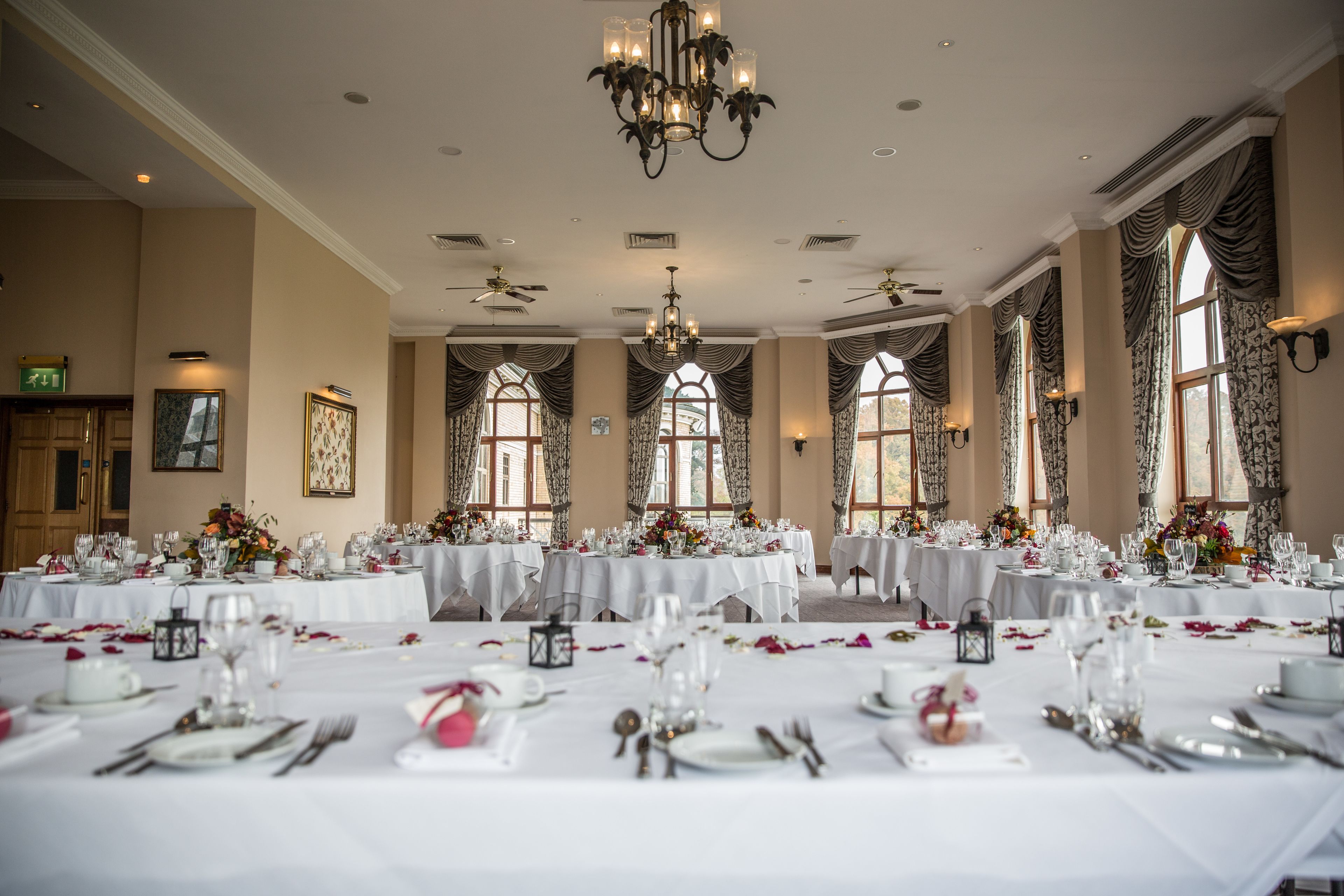 Une salle de banquet avec de nombreuses tables rondes dressées pour un événement formel, de grandes fenêtres ornées de rideaux et un lustre.