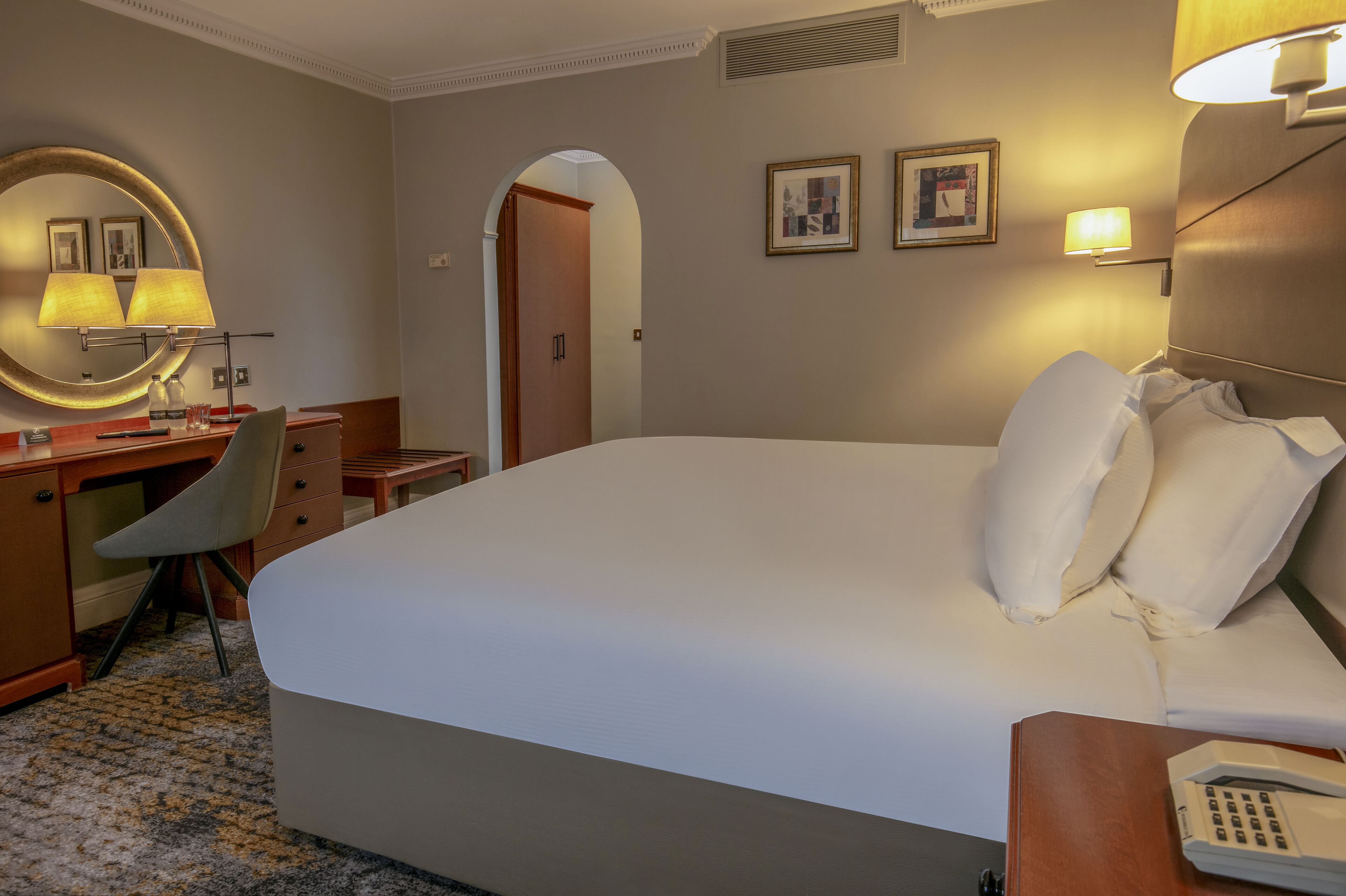 A hotel room with a large bed, a desk with a chair, a round mirror, and two wall-mounted lamps.