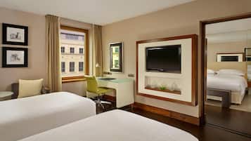 Habitación de hotel con dos camas, un escritorio, un televisor y una ventana que ofrece vistas a edificios.