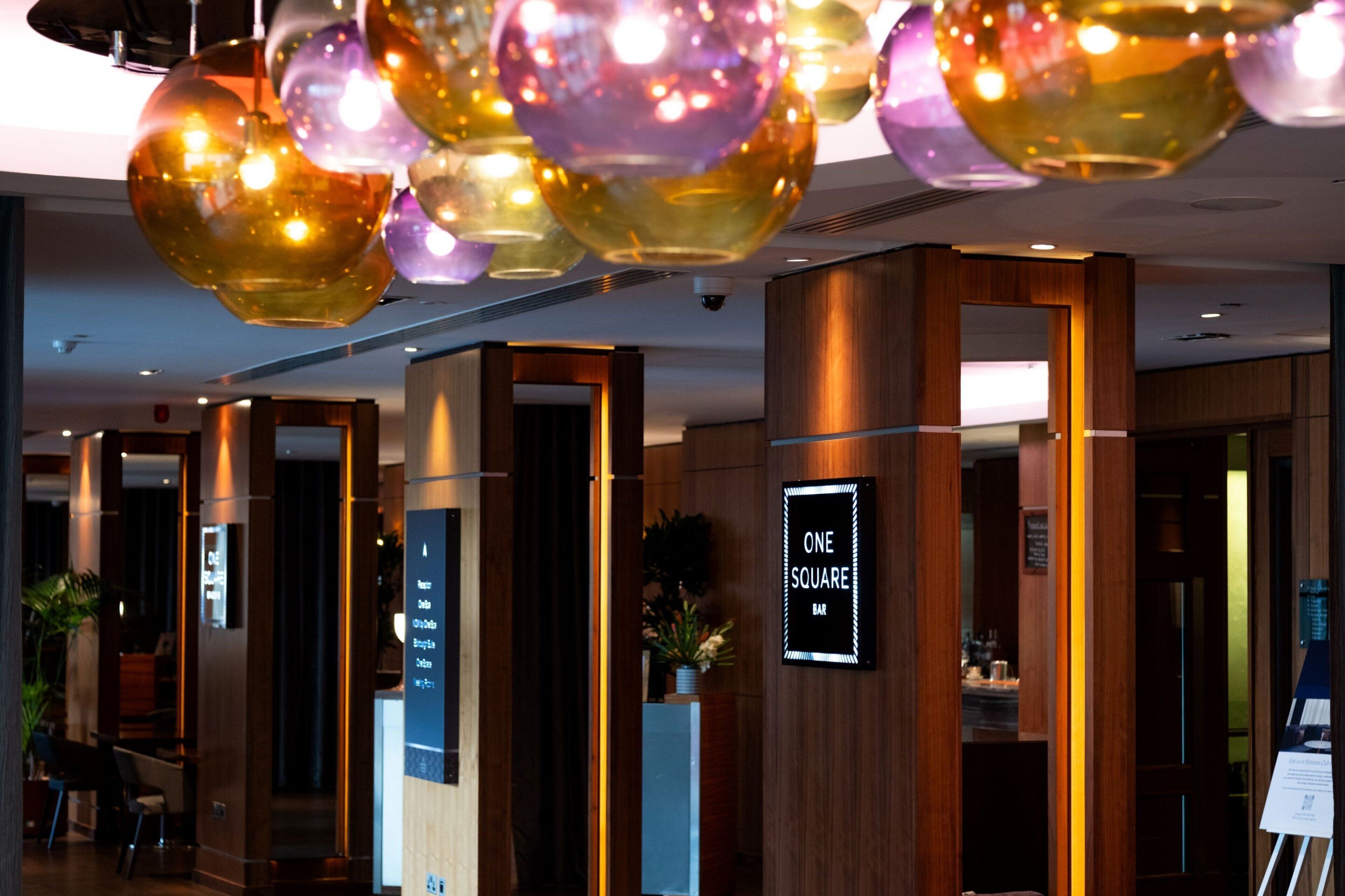 A hotel lobby with a reception desk, a sign for 'ONE SQUARE BAR', and a modern chandelier.