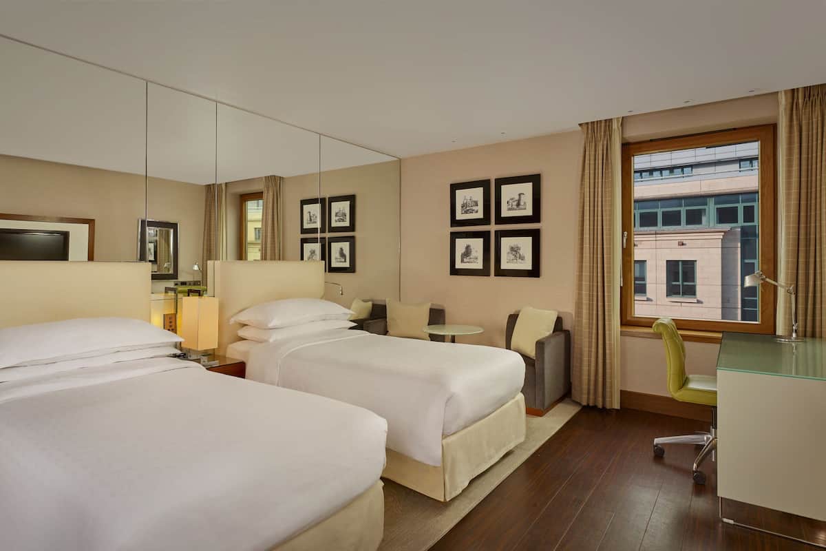 A hotel room with two beds, a desk, a chair, a small table, and a window with curtains.