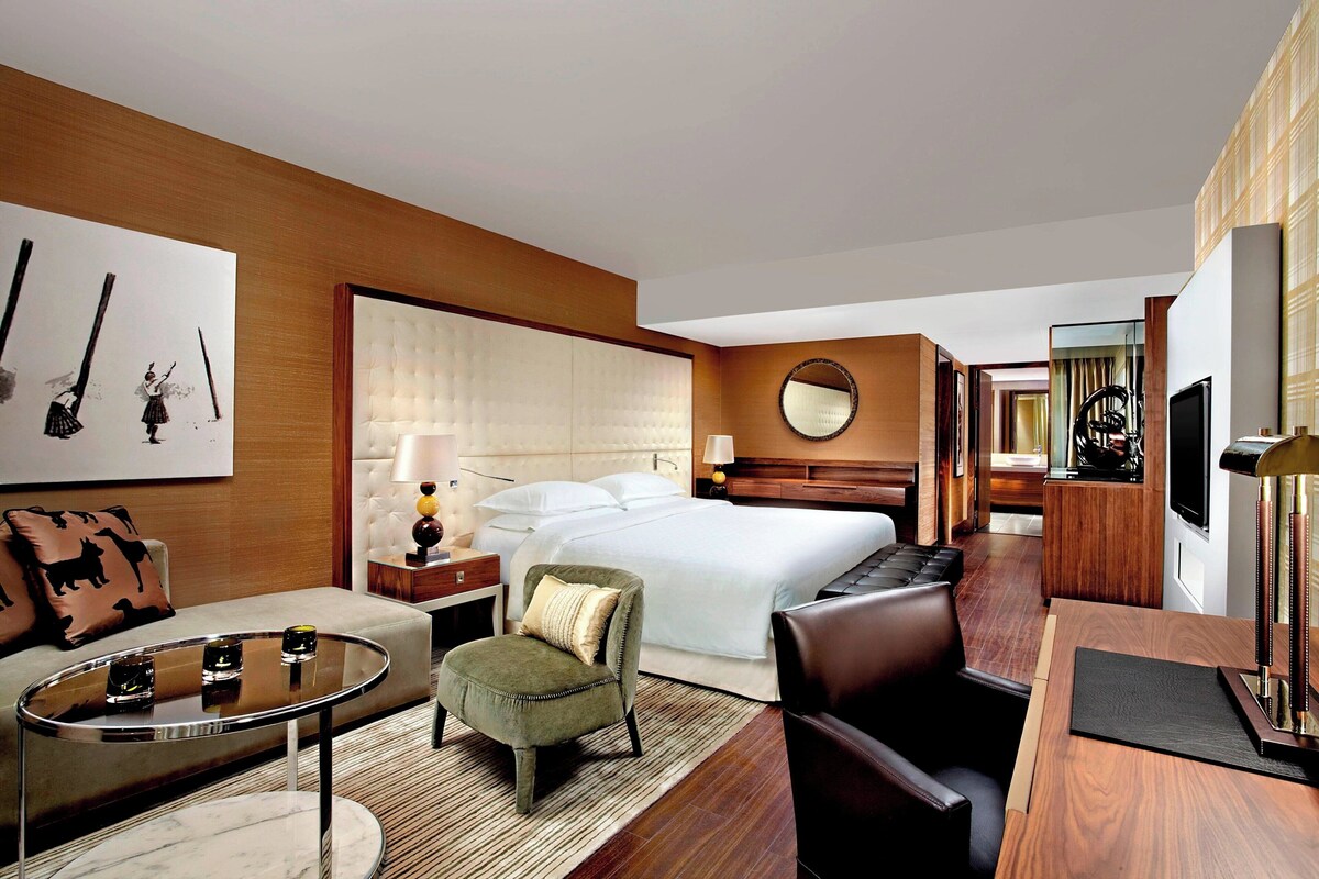 Una habitación de hotel con cama, una zona de estar con mesa y sillas, un televisor y un espejo.