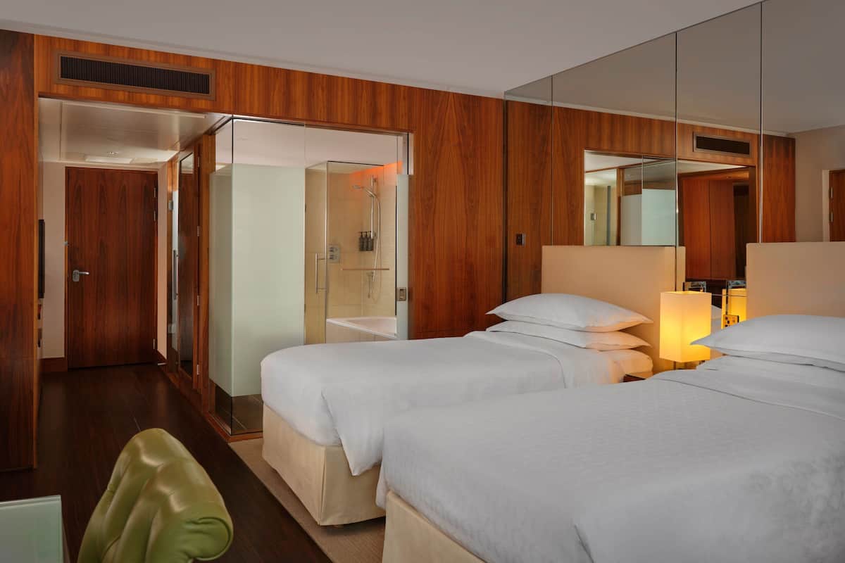 A hotel room with two beds, a mirror, a bedside table with a lamp, and a shower area.