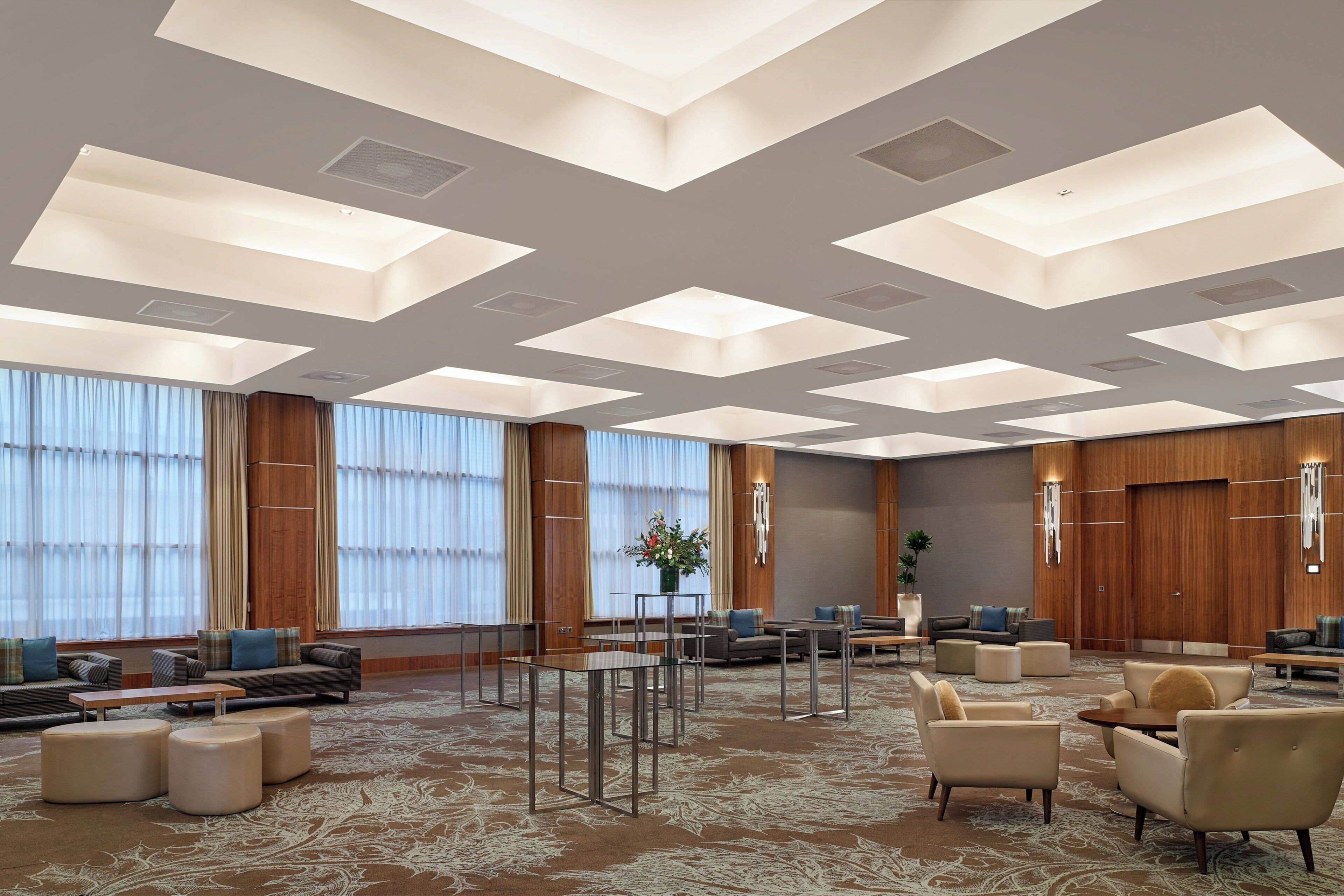 Een ruime lobby met een hoge plafond, grote ramen en diverse zithoekjes, waaronder banken en fauteuils.