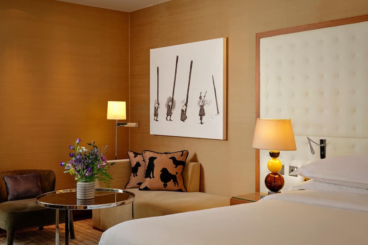 A hotel room with a bed, a sofa, a small table with a vase of flowers, a lamp, and a wall-mounted artwork.