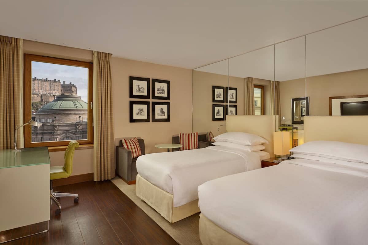 A hotel room with two beds, a desk, a chair, a small table, and a large window with a view of a historic building.