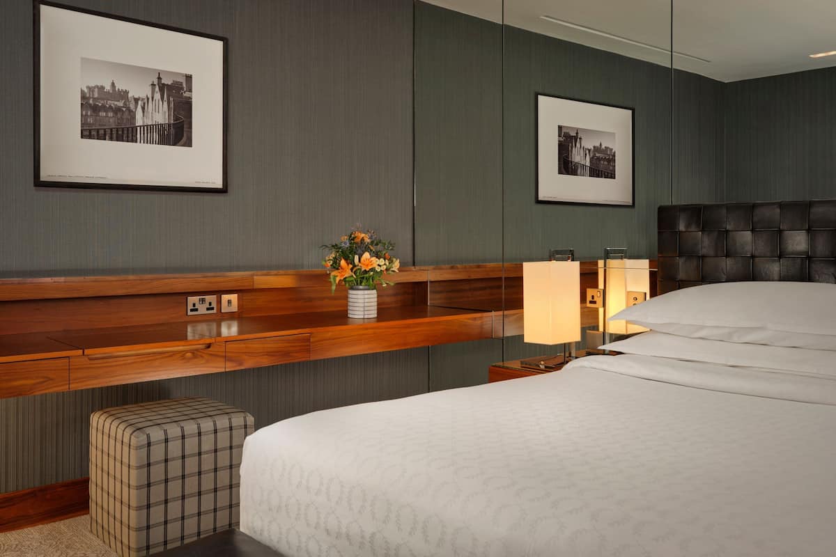 A hotel room with a large bed, a wooden console table, a small stool, a nightstand with a lamp, and two framed pictures on the wall.