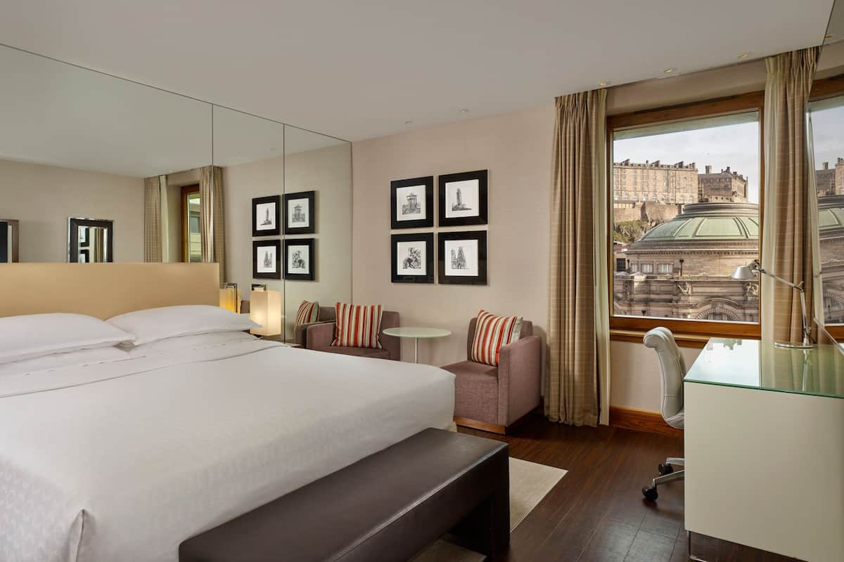 A hotel room with a large bed, a desk, a chair, a small table, and a large window with a view of buildings.