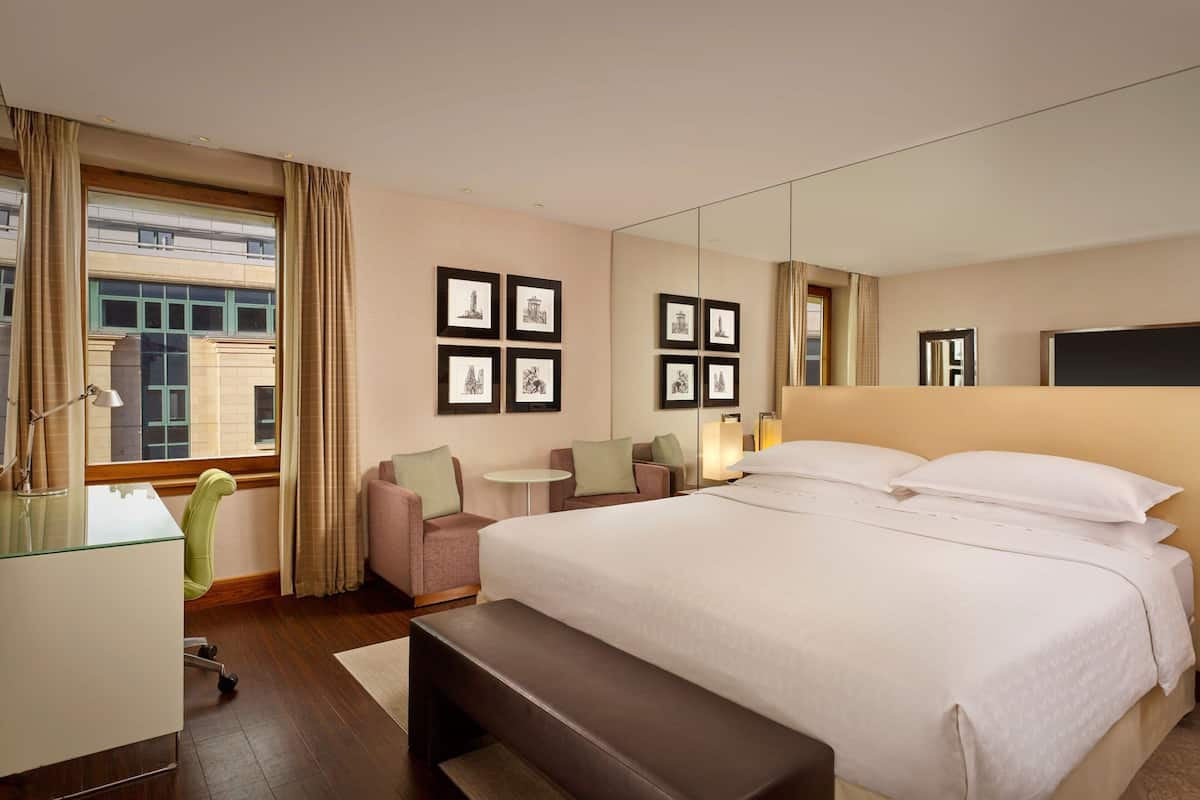 A hotel room with a large bed, a desk, a chair, a small table, and a bench.