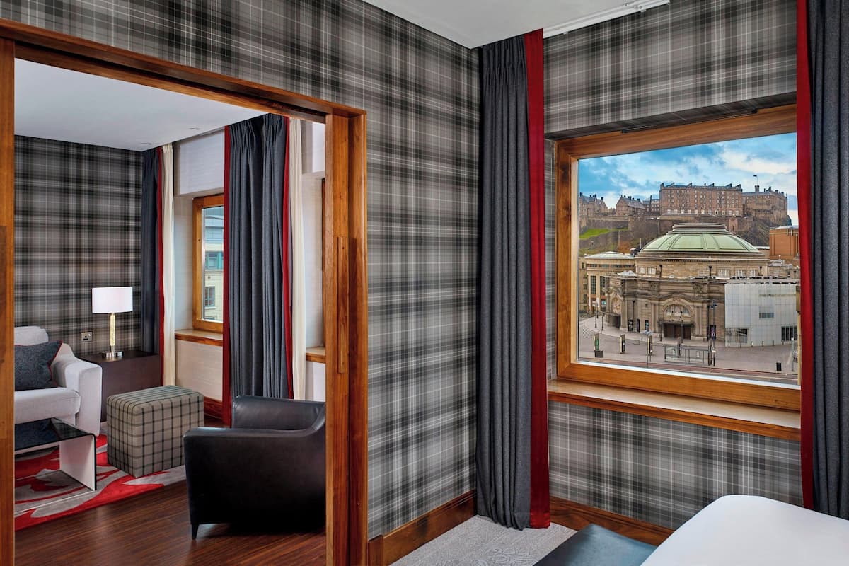 A hotel room with a plaid wallpaper, a large window with a view of a historic building, a sofa, a chair, and a small table.