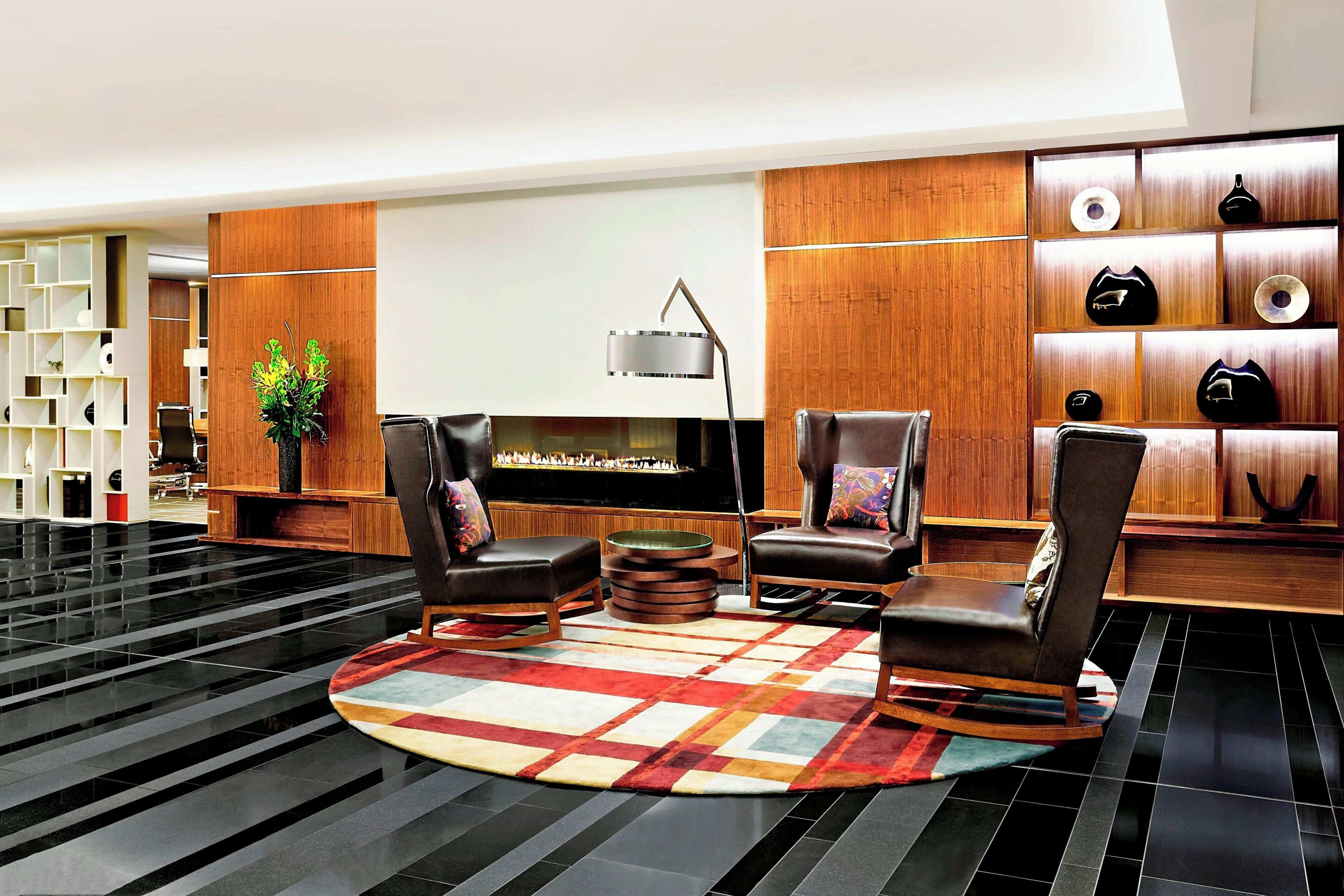 A modern lobby with leather chairs, a round coffee table, a patterned rug, a bookshelf, and a wall-mounted shelf with decorative items.