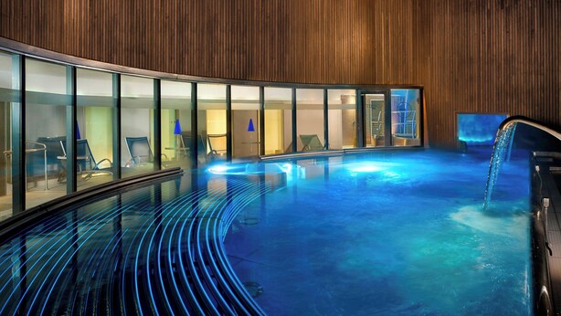 A modern indoor pool with a curved glass wall, wooden paneling, and a blue-lit floor.