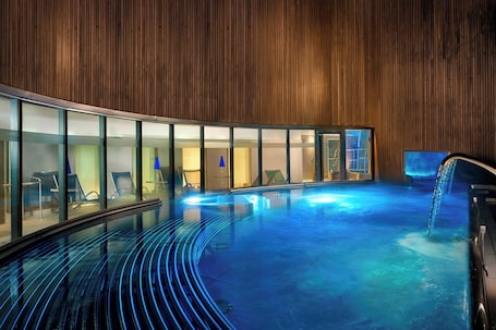 A modern indoor pool with a curved glass wall, wooden paneling, and a blue-lit floor.