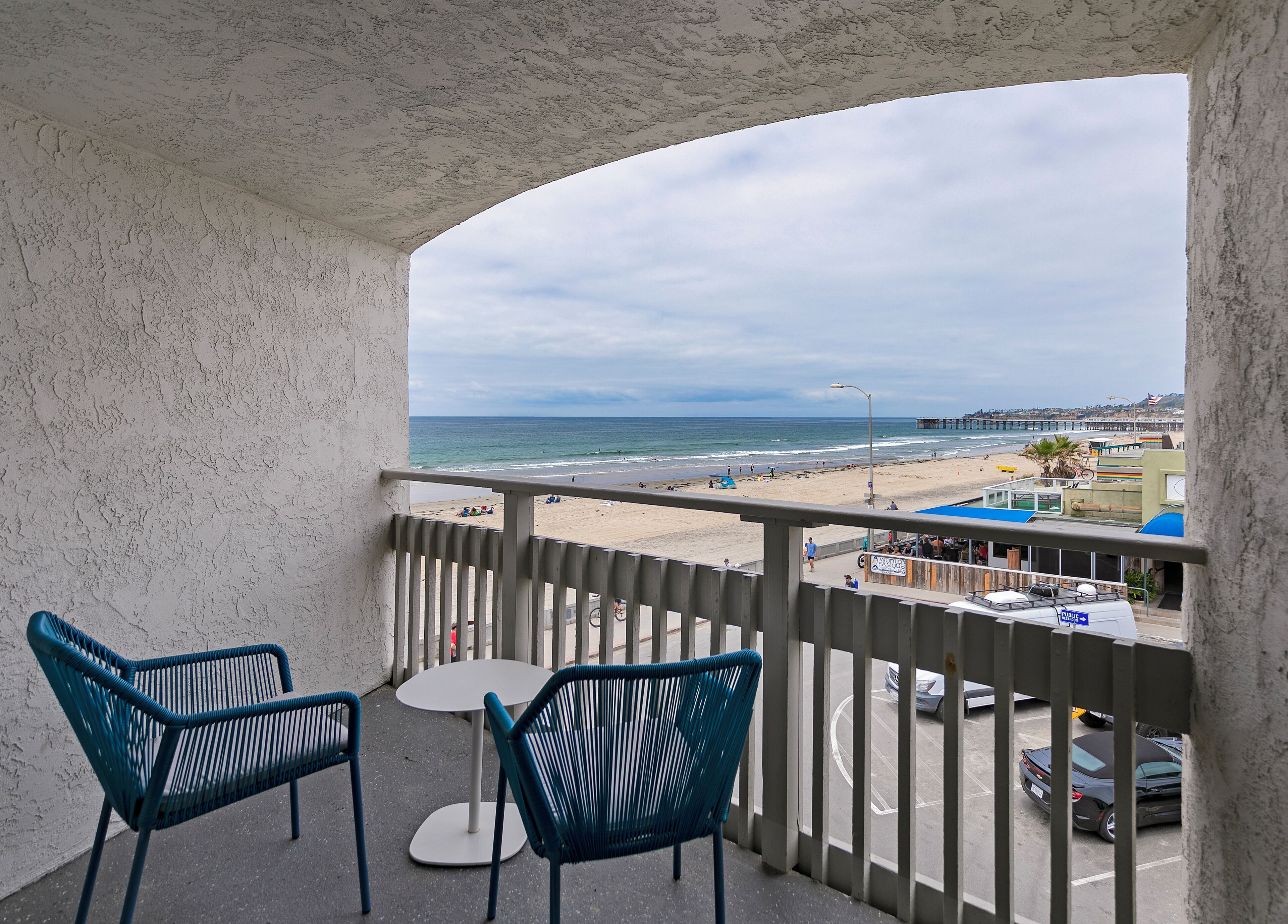 Un balcón con dos sillas y una mesa pequeña, con vistas a una playa que tiene un muelle y un aparcamiento.