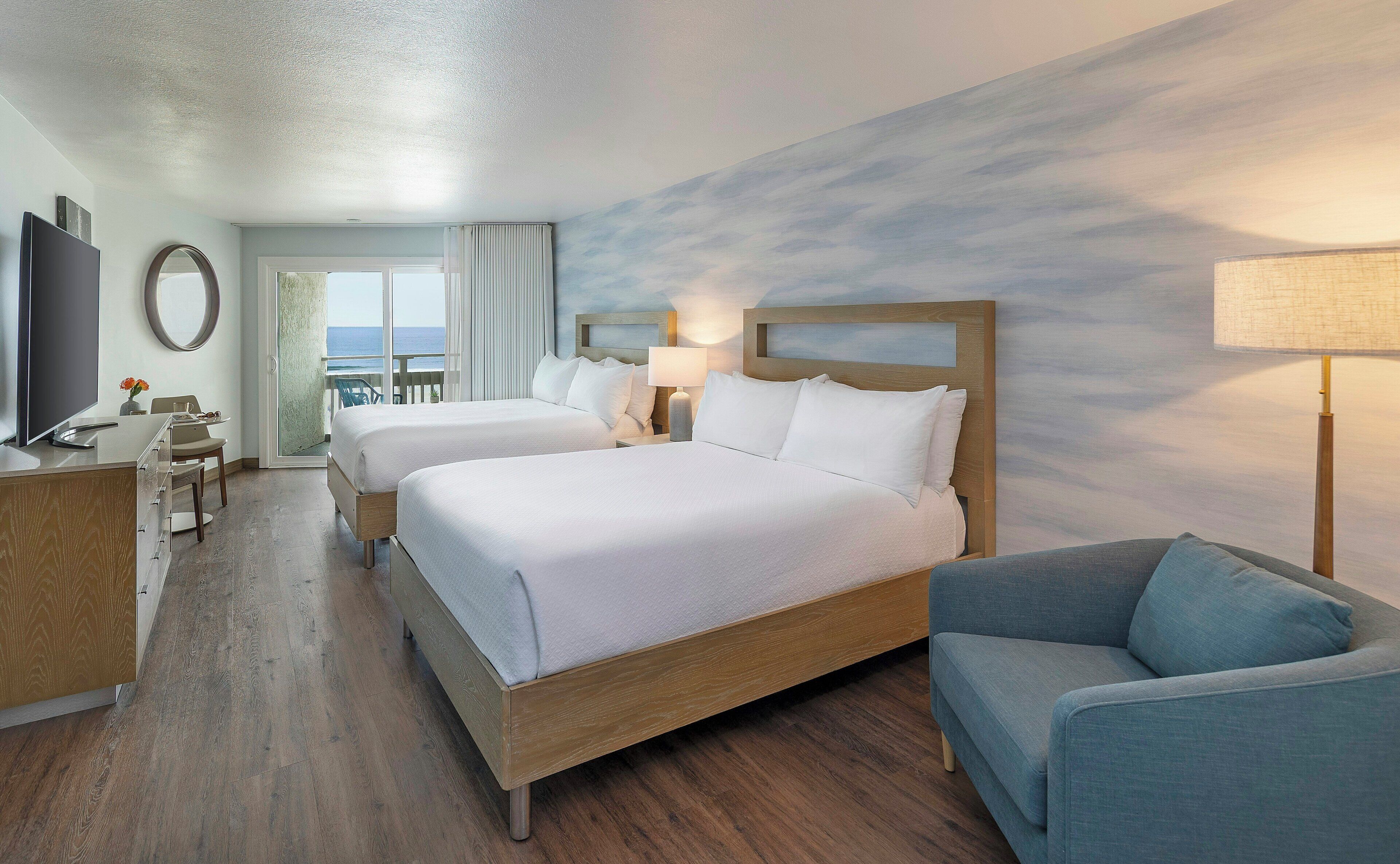 A hotel room with two beds, a TV, a desk, a chair, and a view of the ocean.