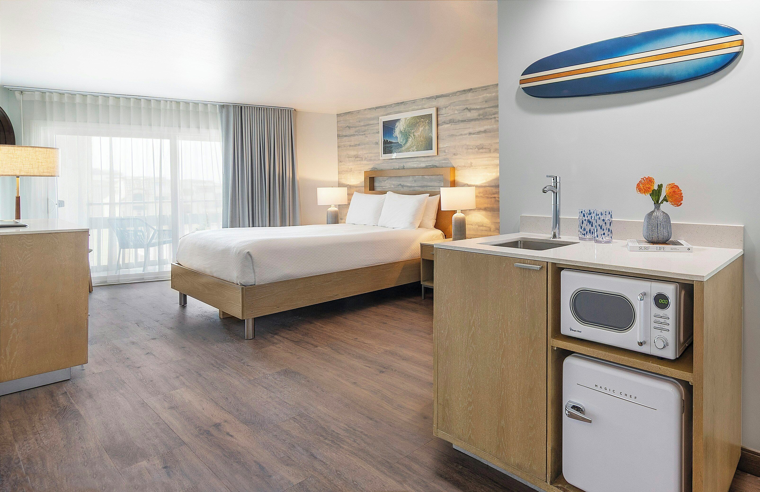 A hotel room with a bed, bedside lamps, a microwave, a refrigerator, a sink, and a window with curtains.