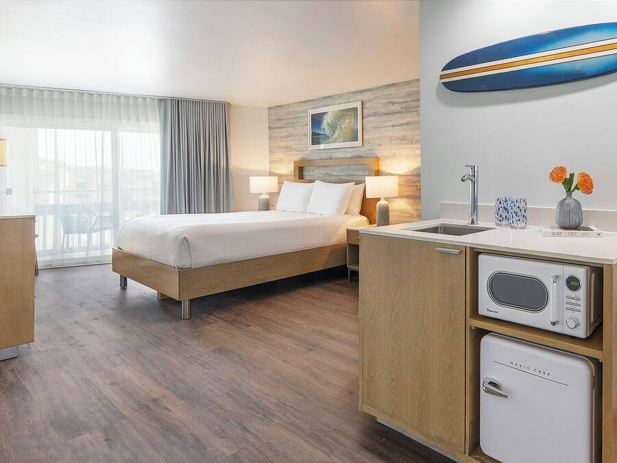A hotel room with a bed, bedside lamps, a microwave, a refrigerator, a sink, and a window with curtains.