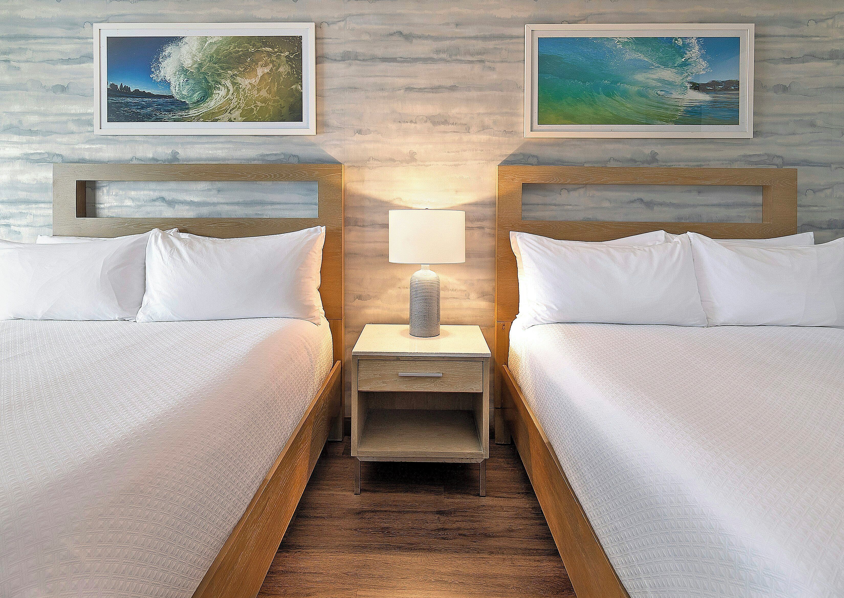 A hotel room with two beds, a nightstand with a lamp, and two framed pictures on the wall.