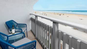 A balcony with blue chairs and a table, overlooking a beach with people and umbrellas.