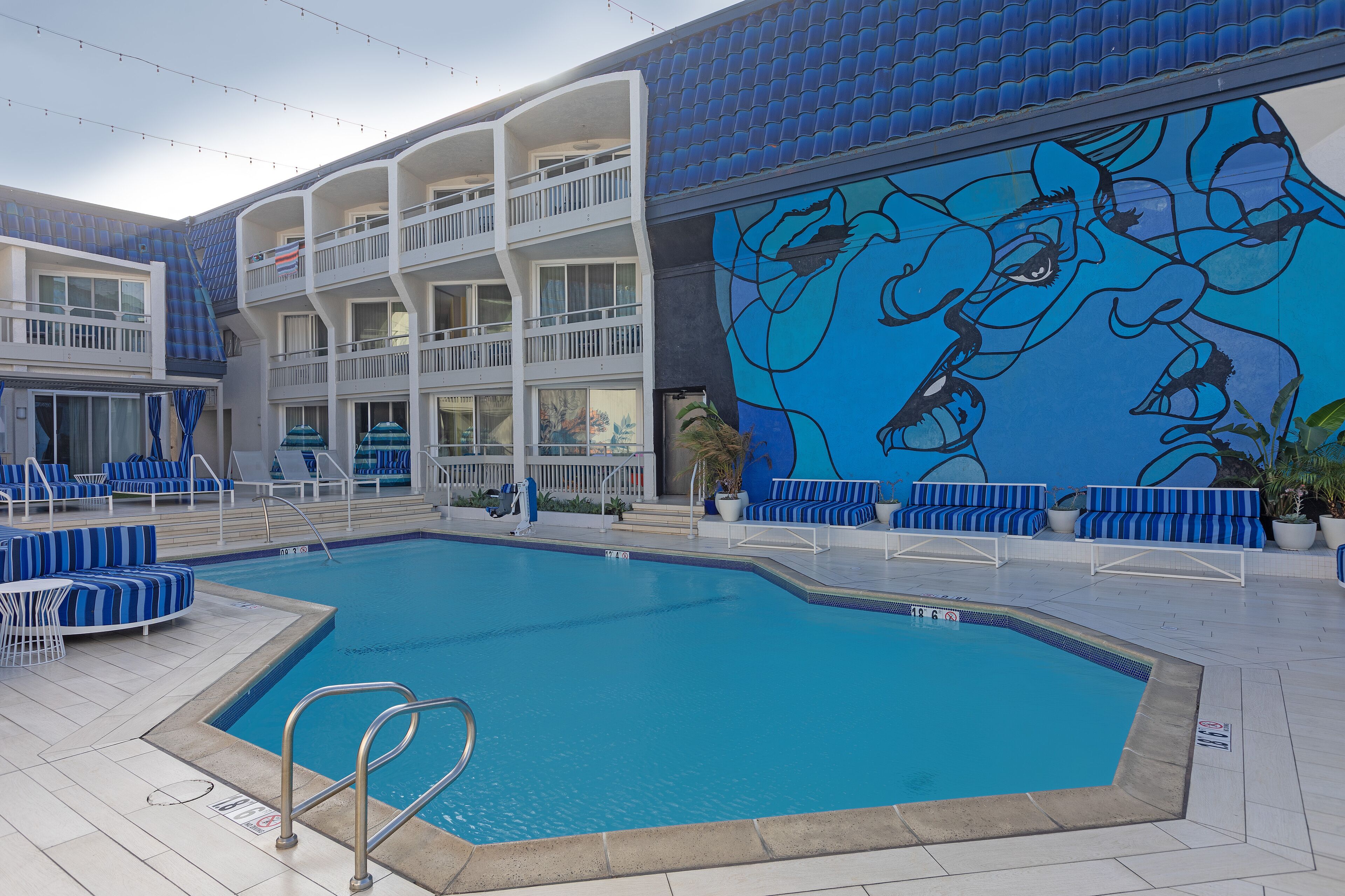 Una piscina con sillas reclinables azules y blancas, un mural y un edificio de varios niveles con balcones.
