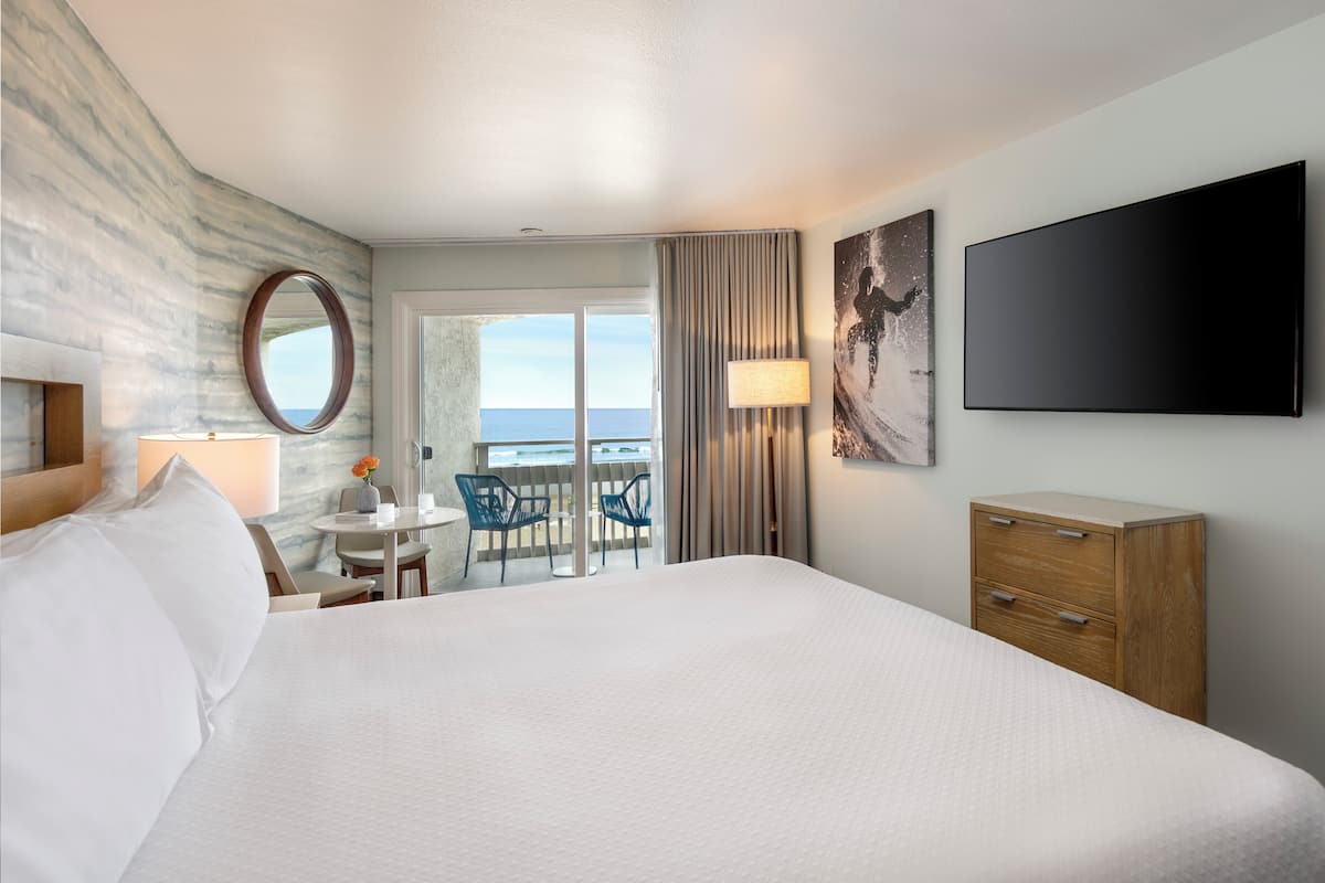 Une chambre d’hôtel avec un grand lit, un bureau avec des chaises, une télévision et une vue sur l’océan.