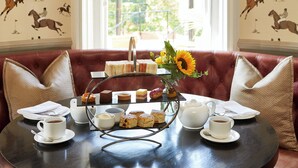 A dining table with a tea set, sandwiches, and pastries, a vase with sunflowers, and a red upholstered bench. - Macdonald Berystede Hotel & Spa (Ascot)