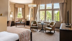 A hotel room with a bed, two armchairs, a round table, a television, and large windows with curtains. - Macdonald Berystede Hotel & Spa (Ascot)