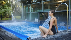 A woman sitting by a hot tub with a waterfall feature, surrounded by a modern building with large windows. - Macdonald Berystede Hotel & Spa (Ascot)