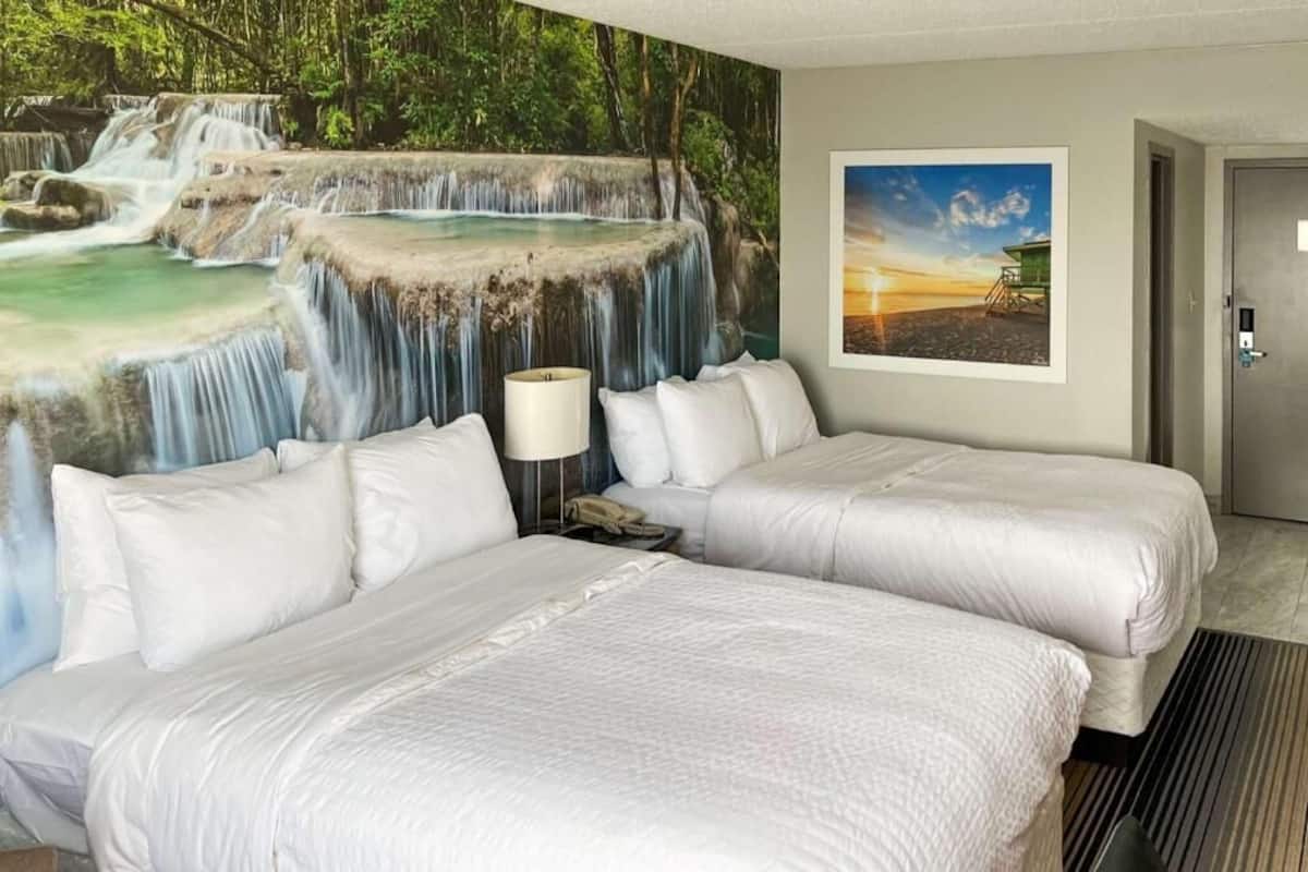A hotel room with two beds, a mural of a waterfall, a framed sunset photo, and a door.