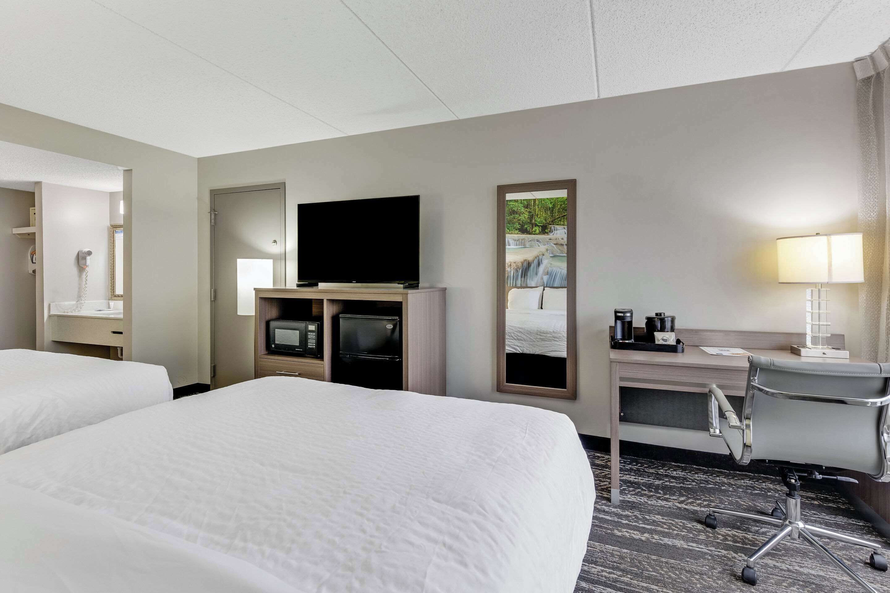 Ein Hotelzimmer mit zwei Betten, einem Schreibtisch mit Computer, einem Flachbildfernseher, einer Lampe und einem Spiegel.