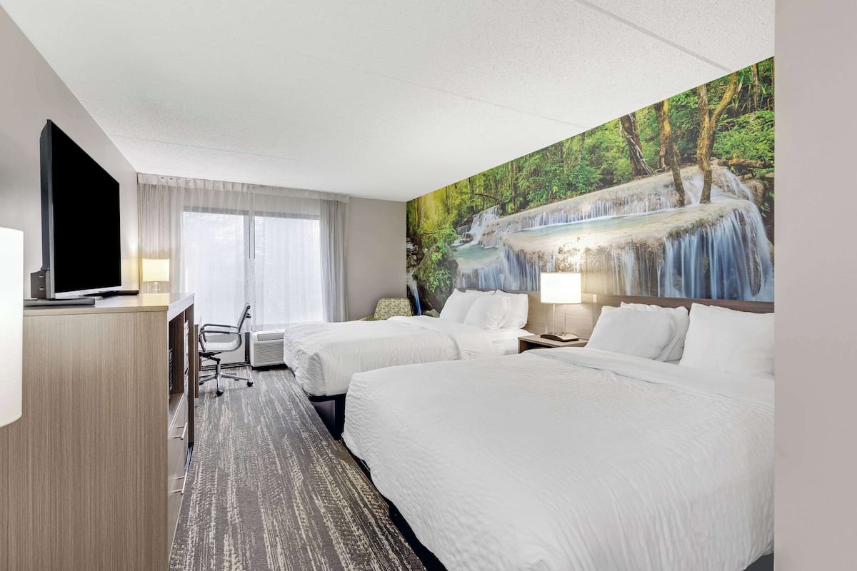 A hotel room with two beds, a desk, a TV, and a mural of a waterfall.