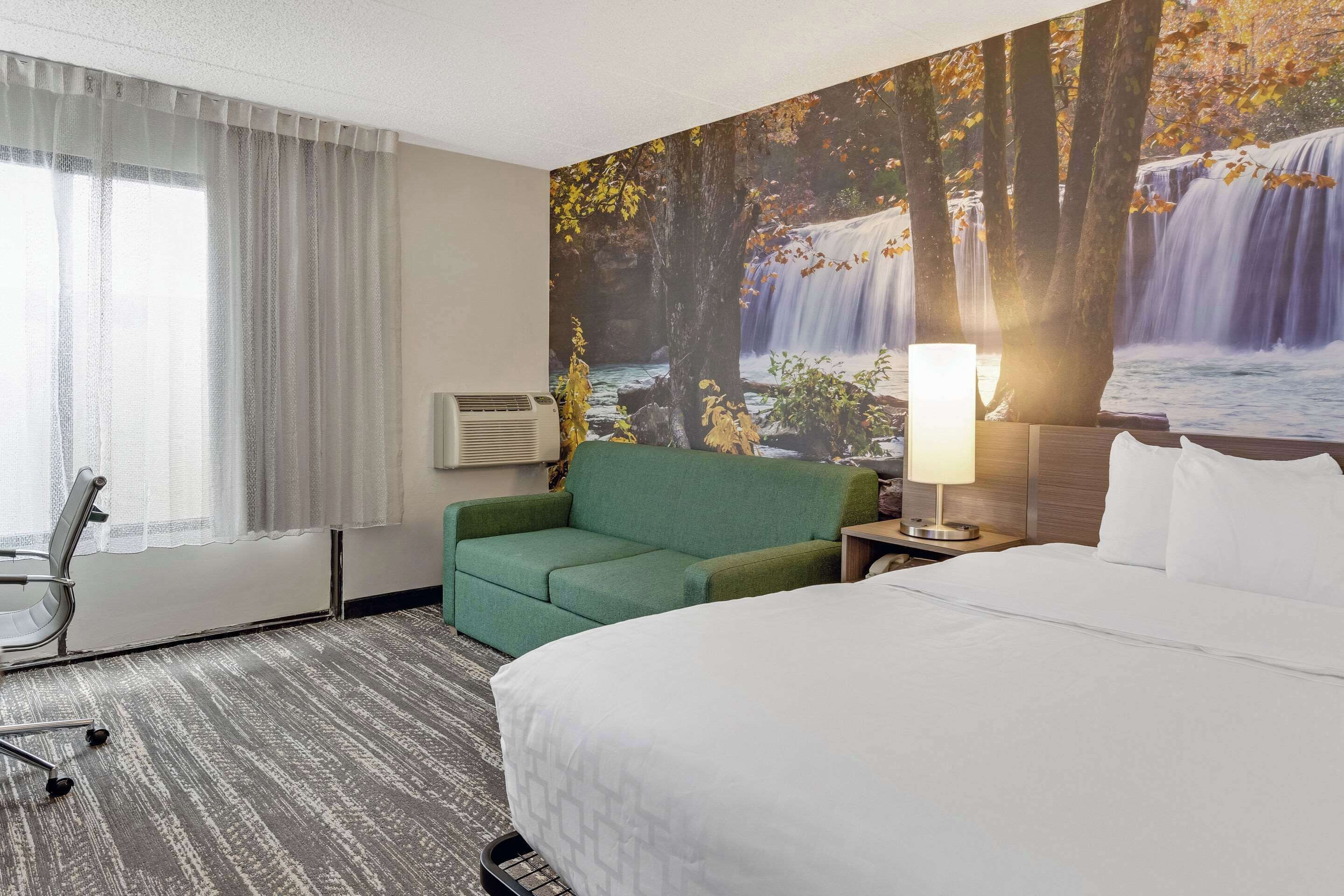 A hotel room with a bed, a green sofa, a desk with a lamp, and a large wall mural of a waterfall.