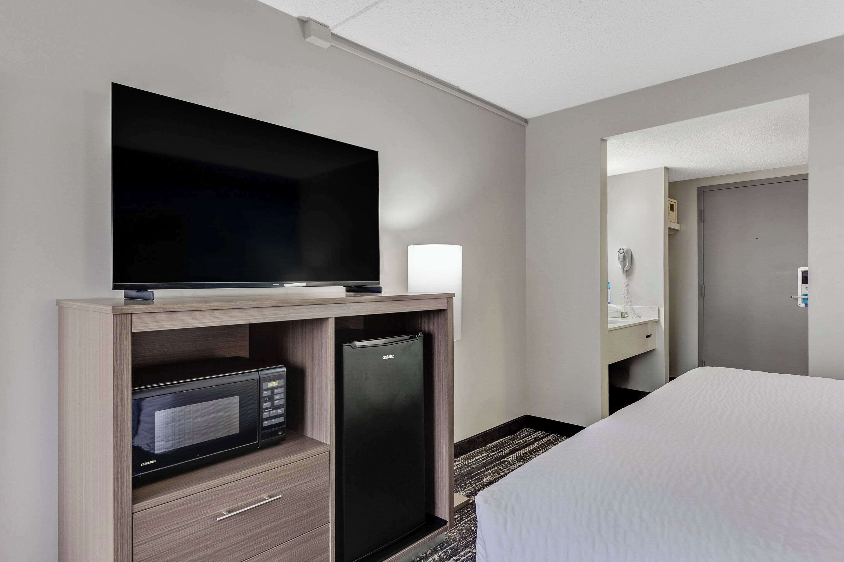 A hotel room with a flat-screen TV on a wooden stand, a minibar with a microwave, a refrigerator, a bed with white bedding, a bathroom with a mirror and sink, and a door.