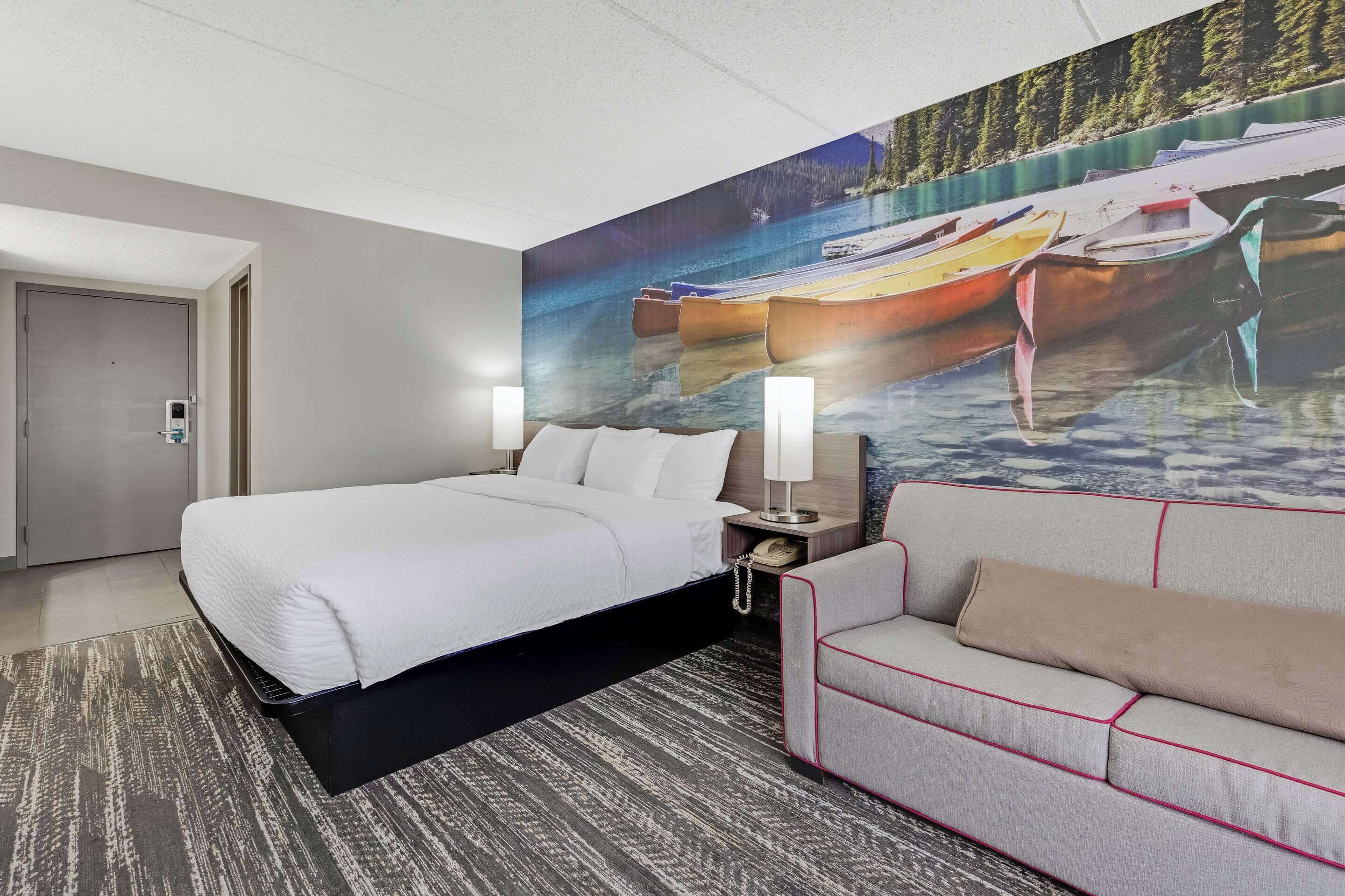 A hotel room with a large bed, a sofa, a nightstand with a lamp, and a mural of boats on water.