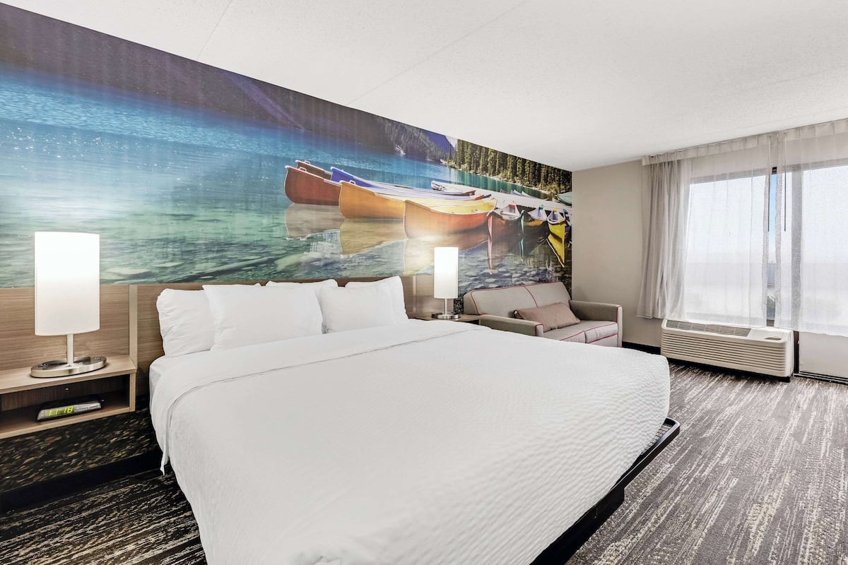A hotel room with a large bed, a desk with a lamp, a chair, and a large window with curtains.