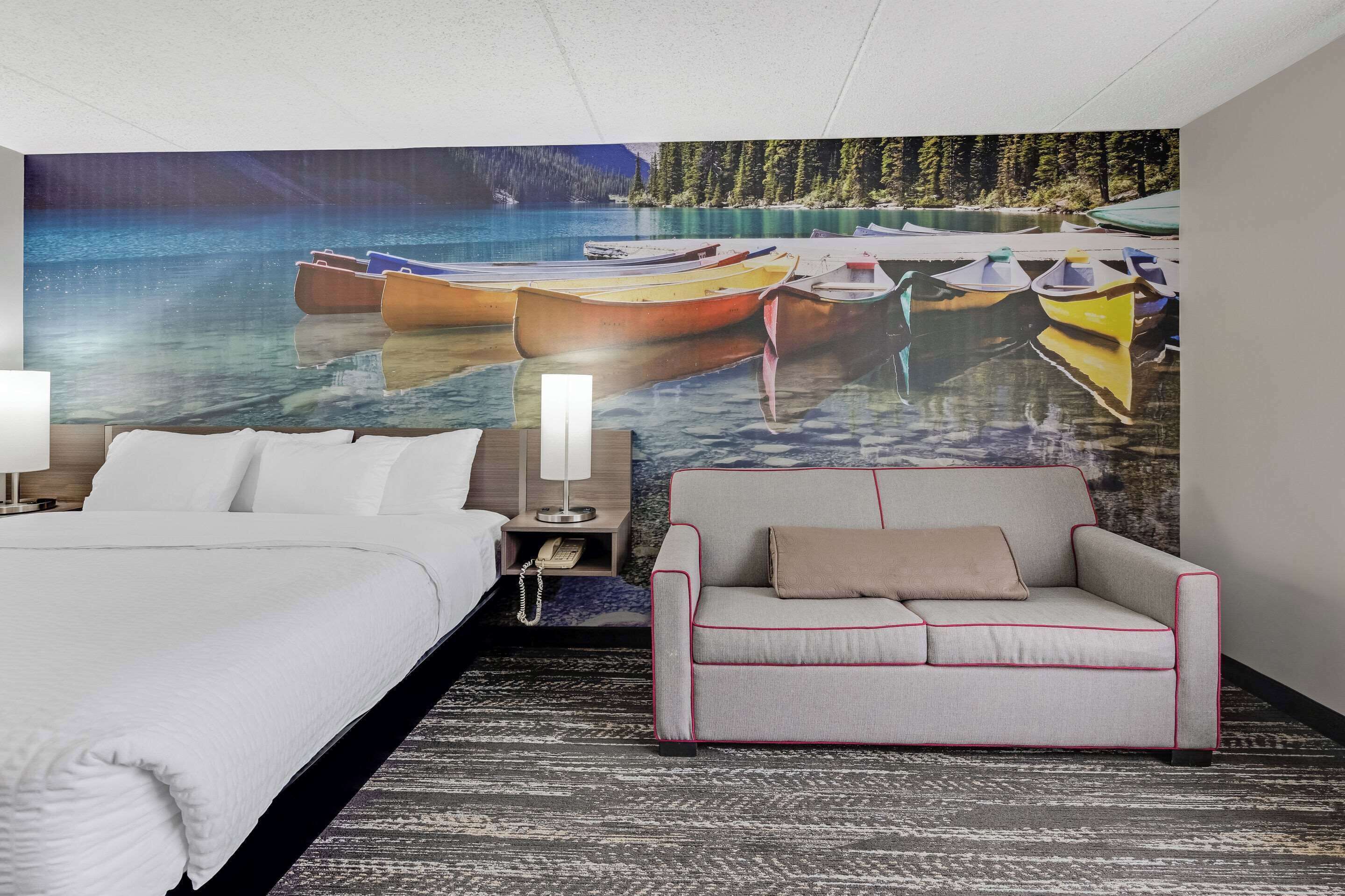 Ein Hotelzimmer mit Bett, einer Couch, einem Nachttisch mit Lampe und einem großen Wandbild von Booten auf dem Wasser.