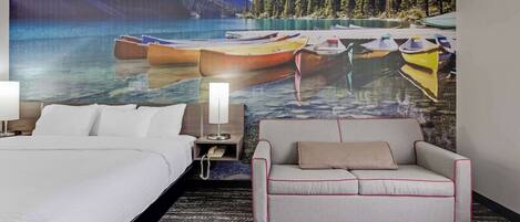 A hotel room with a bed, a sofa, a nightstand with a lamp, and a large wall mural of boats on water.