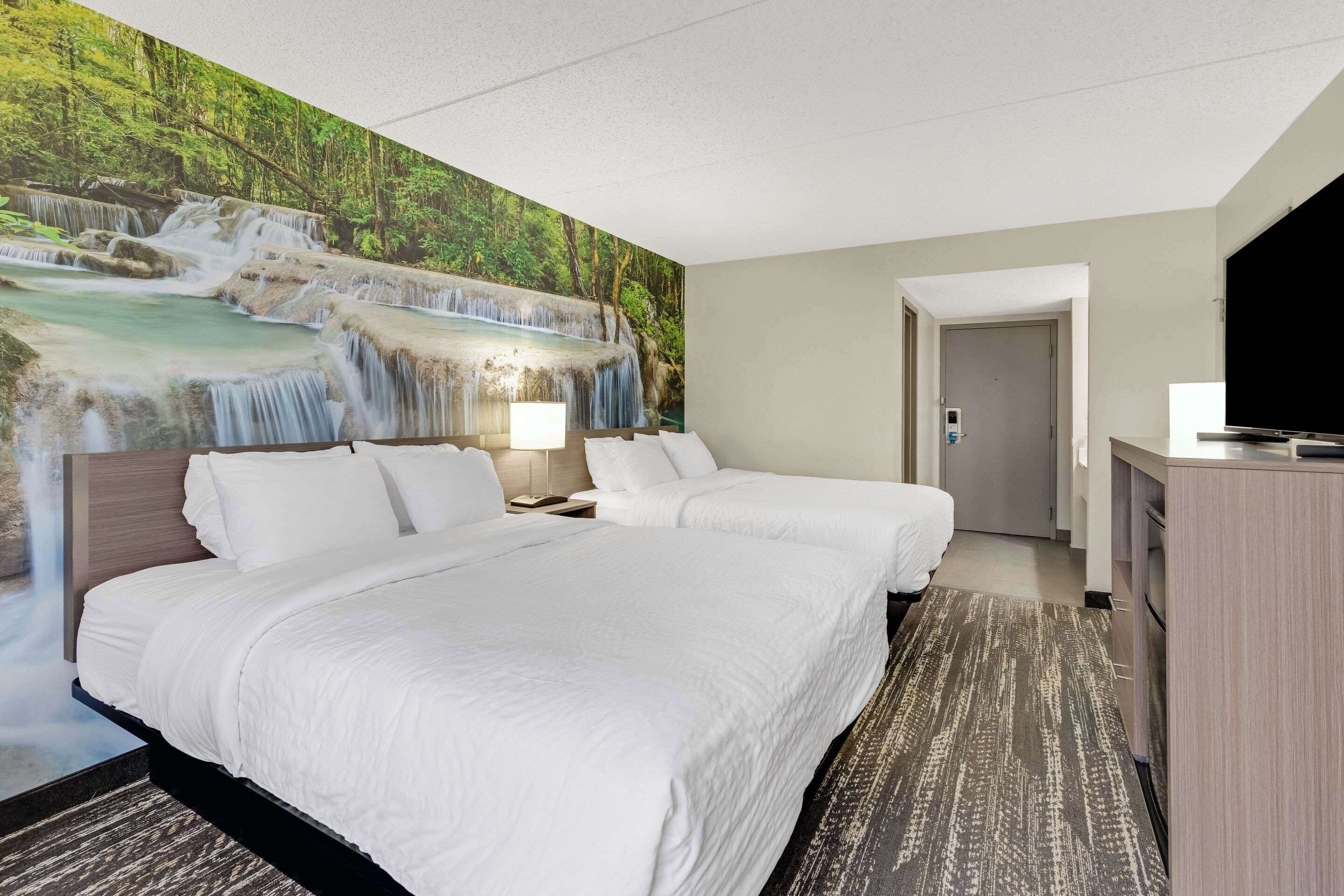 A hotel room with two beds, a large mural of a waterfall, a desk with a TV, and a door leading to a bathroom.