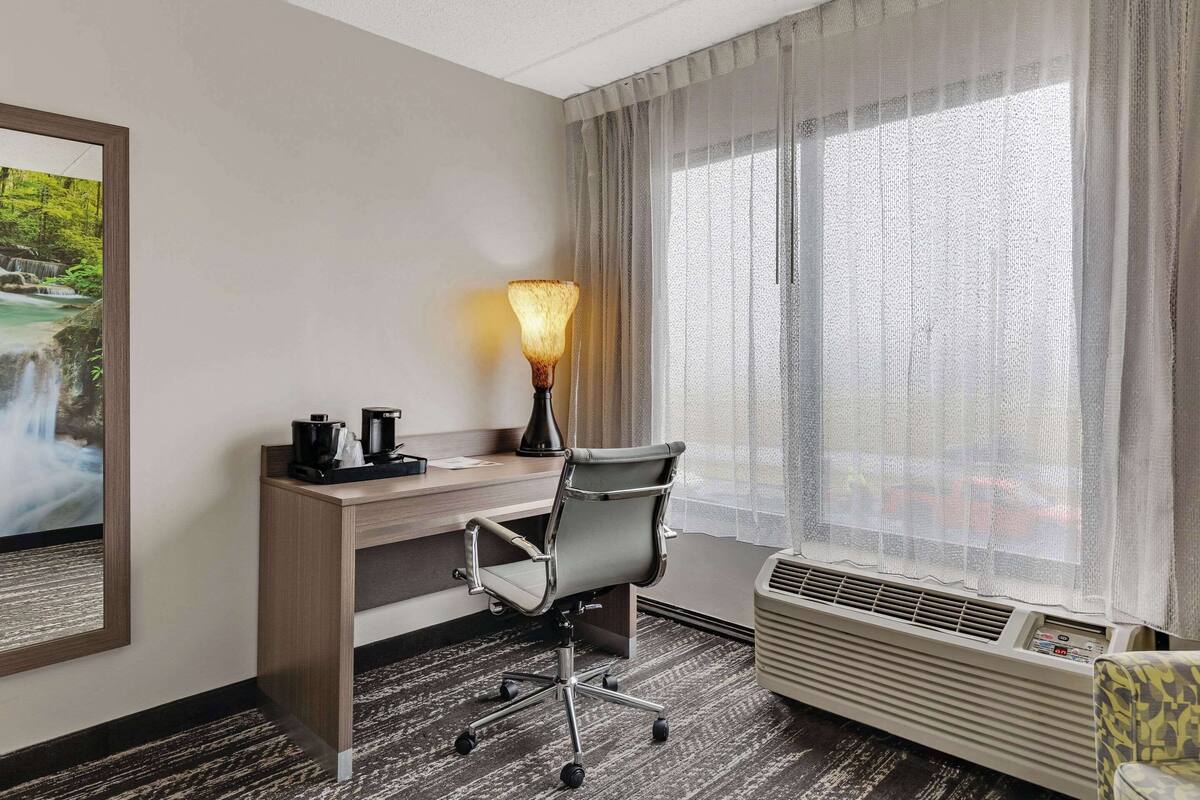 A hotel room with a desk, chair, lamp, air conditioner, and a large window with sheer curtains.