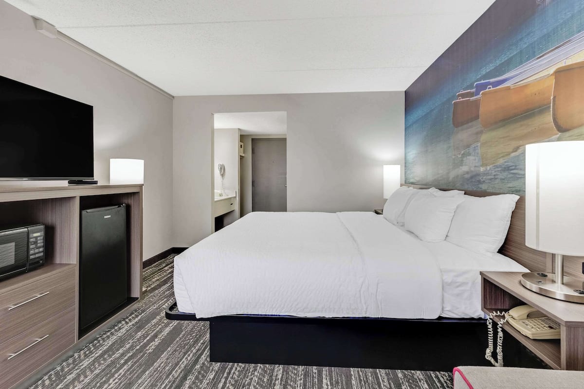 A hotel room with a bed, a television, a refrigerator, a desk with a lamp, and a painting on the wall.